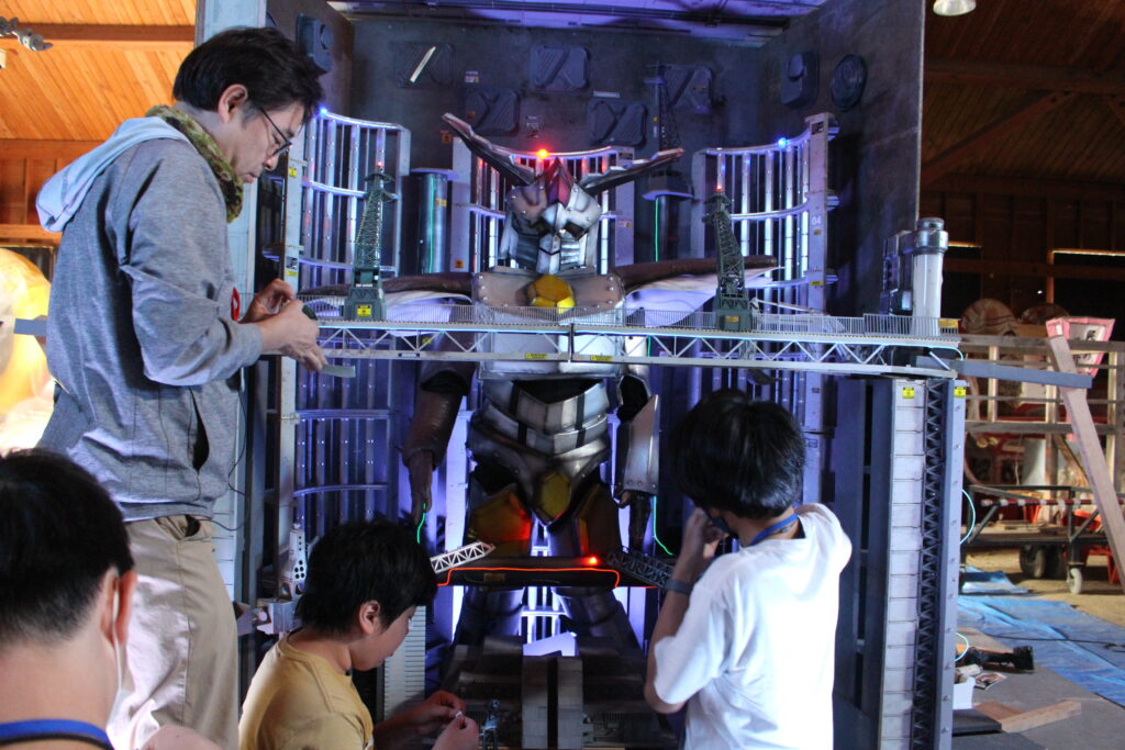 巨大ロボット模型の前で複数の人物が周囲の装置やパーツを調整し発光演出の状態を確認しながら作業を進める様子の写真