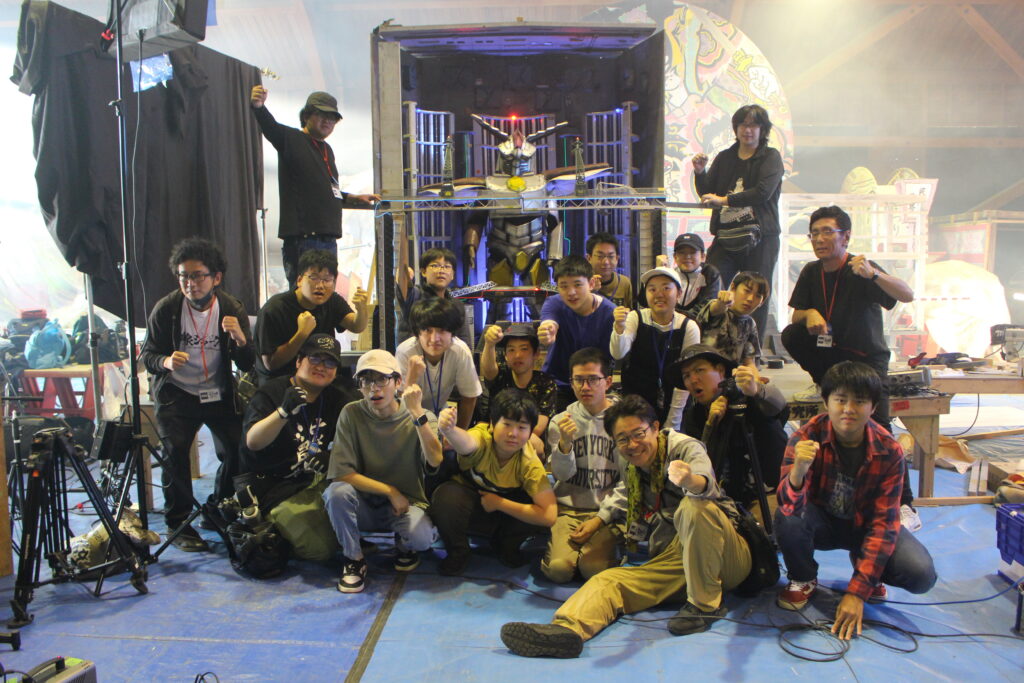 多数の参加者が巨大ロボット模型の前に集まり拳を掲げて統一ポーズを決めながら記念撮影に臨む様子の写真