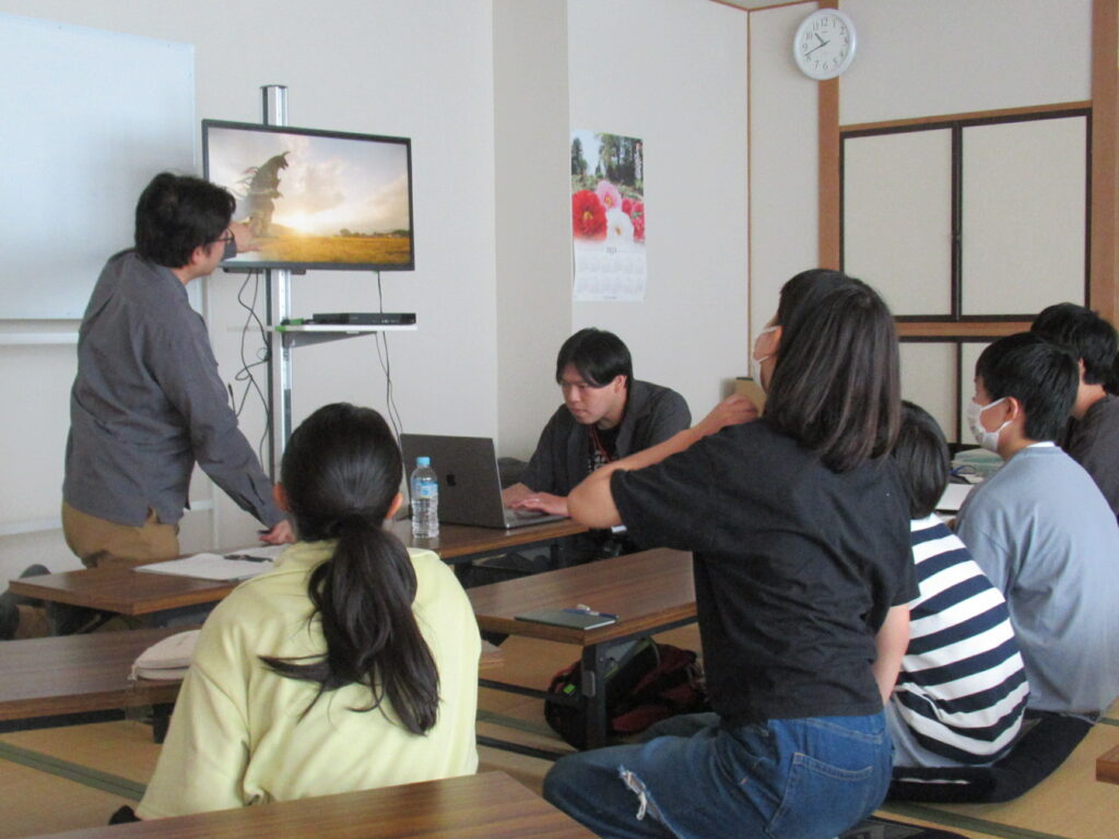 明るい室内で講師がモニターに映る怪獣の映像を指差しながら解説し、それを見つめる数名の塾生が畳の上に座って熱心に耳を傾けている講習会の様子の写真