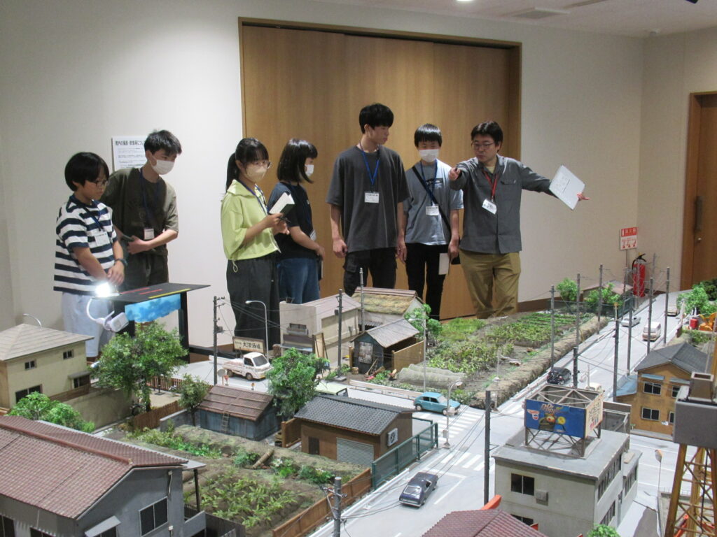 屋内の広い展示スペースで、講師が指を差しながら解説を行い、その横で数名の塾生が立ち並んで精巧に作られた住宅街や道路、畑などの大規模なジオラマを熱心に見学している写真