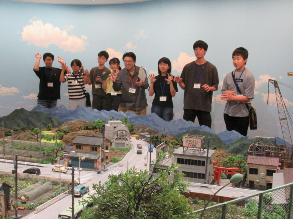 精巧に作られた山々や街並みのミニチュアセットを前にして、講師と数名の塾生が怪獣のような手つきでポーズを決めながら、青空の背景パネルを背に和やかな雰囲気の集合写真