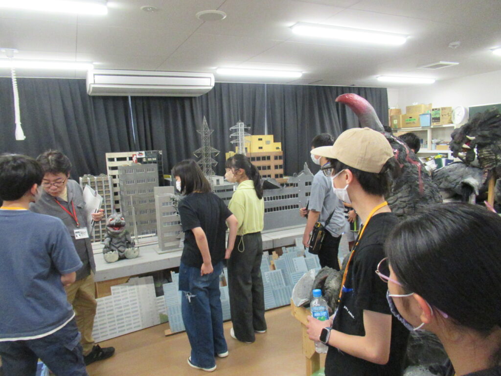 屋内のスタジオで、講師と数名の塾生がミニチュアのビル群や鉄塔、小さな怪獣の模型が置かれた特撮セットを囲み、制作の仕組みや撮影手法について熱心に確認している写真