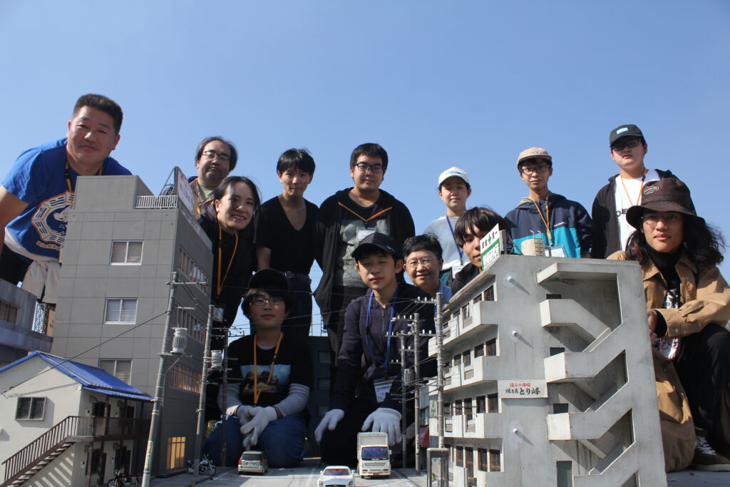 晴れ渡る青空の下で、精巧に作られたビルや住宅、電柱などのミニチュア模型が並ぶ街並みのセットを囲み、制作に携わった多くのスタッフや塾生の集合写真