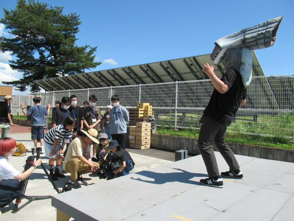 屋外の特撮セットで、大きな怪獣の被り物をした人物がステージ上でポーズを決め、それを講師や塾生がカメラを構えたり見守ったりしながら真剣に撮影の練習を行っている写真