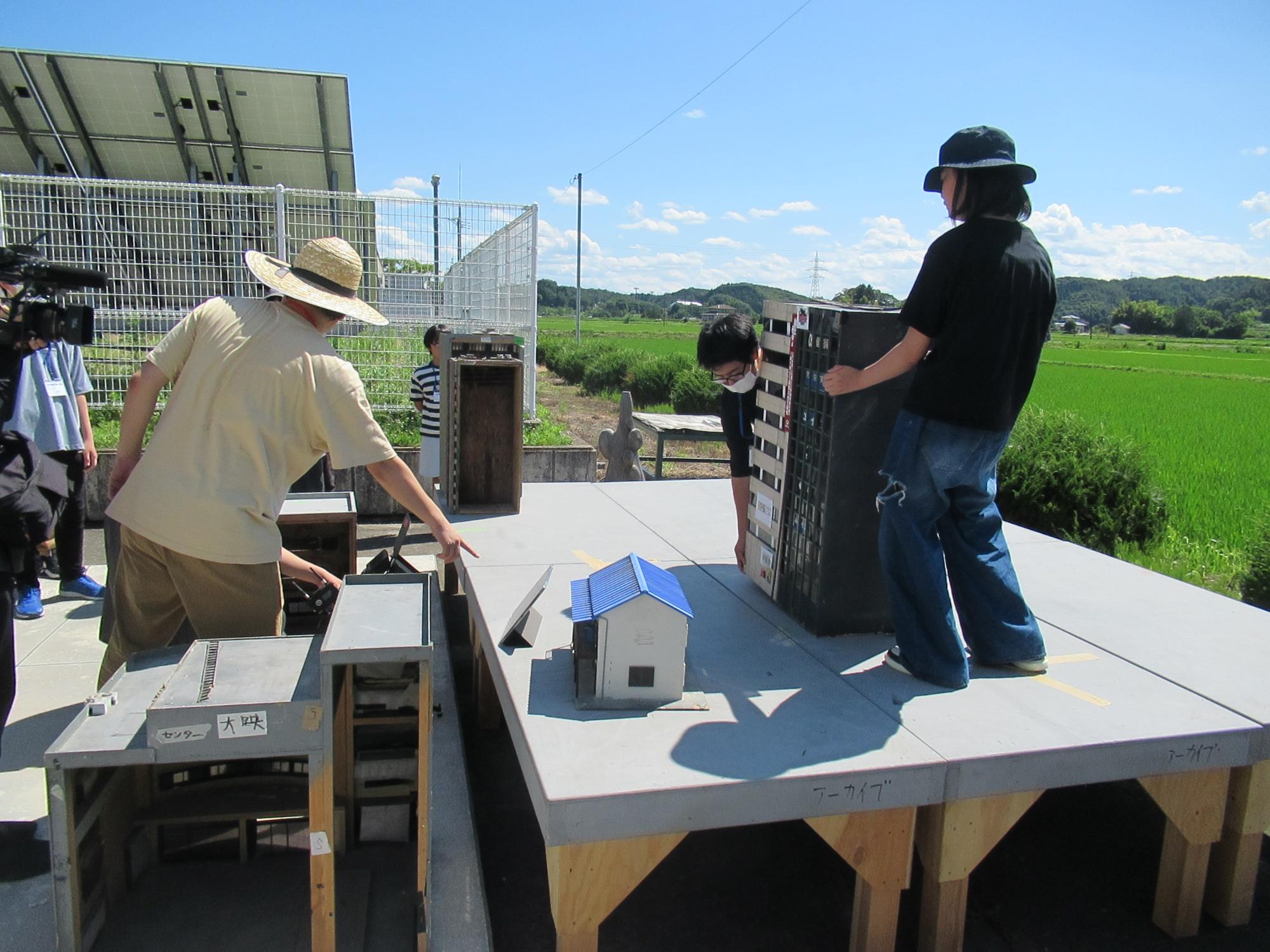 すかがわ特撮塾の活動で模擬撮影を行っている様子、ミニチュアセットをくみ上げるために、2人の学生が監督の指示に従って建物のミニチュアを平台の上に置いている