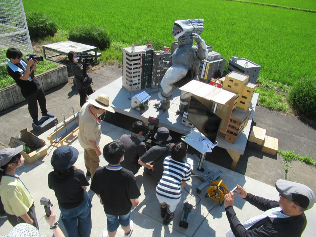 青々とした田んぼを背にミニチュア都市と怪獣模型を囲み撮影機材を操作する参加者たちの様子を収めた写真