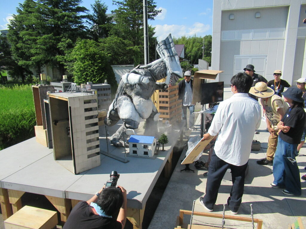 青々とした田園地帯に隣接する屋外セットで、精巧なビル群のミニチュアに囲まれて咆哮するようなポーズをとる巨大な怪獣の着ぐるみを、講師や大勢の塾生がカメラやレフ板を手に取り真剣に撮影している写真