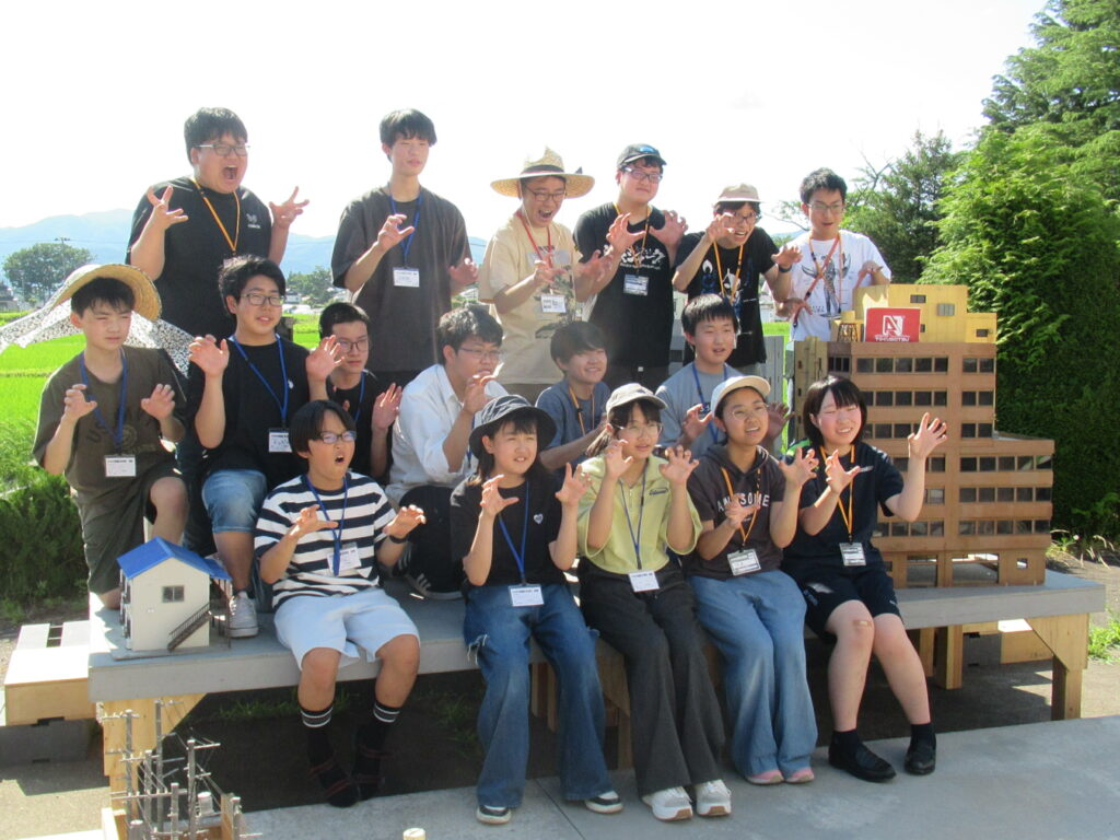 屋外の特撮セットで、精巧なビル群のミニチュアに囲まれながら、怪獣のようなポーズを取り、講師や多くの塾生が和やかな雰囲気で囲んで記念撮影を行っている写真