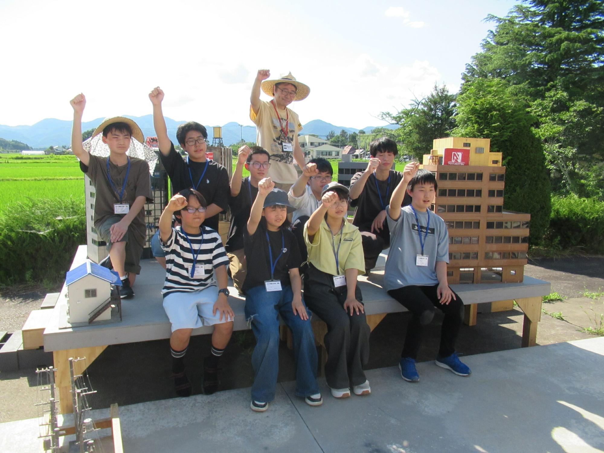 晴天の下、山々を背景にした屋外の特撮セットに集まった10人の塾生が、ミニチュアのビルや建物の模型に囲まれながら、全員で右拳を力強く突き上げてポーズを決めている写真
