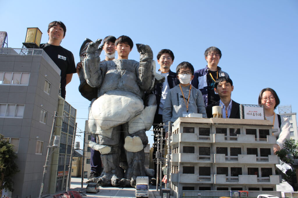 青空が広がる屋外に組まれたビルや住宅の精巧なミニチュア模型の街並みセットを背景に、大きな怪獣の着ぐるみを囲むように複数の塾生が並んで笑顔でポーズを取っている写真