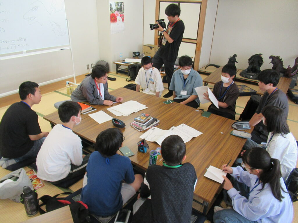 畳の敷かれた和室にて、ホワイトボードに描かれた怪獣のスケッチがあり、その前で講師と数名の塾生が大きな机を囲み座っており、一人の男の子が資料を手に持ち発表している写真