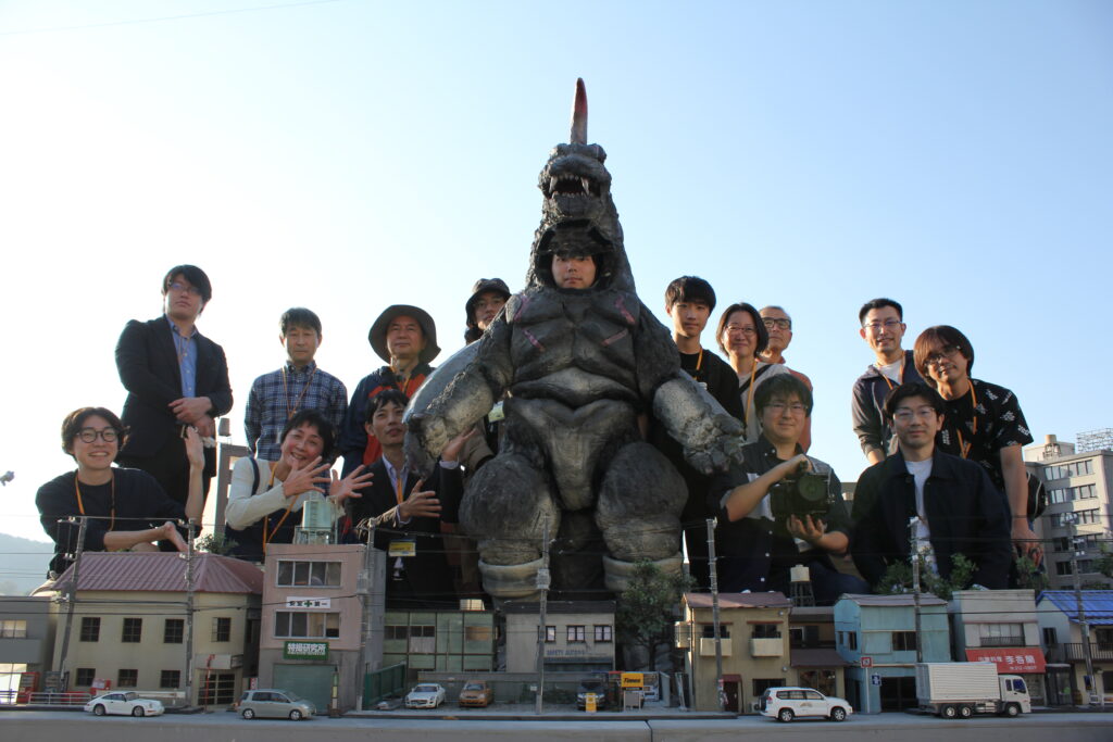 明るい陽光の下、建物や電柱、車などが細部まで再現された精巧なミニチュアの街並みセットを前に、中央に立つ大きな怪獣の着ぐるみを囲んで大勢の塾生が笑顔やポーズで並んでいる写真