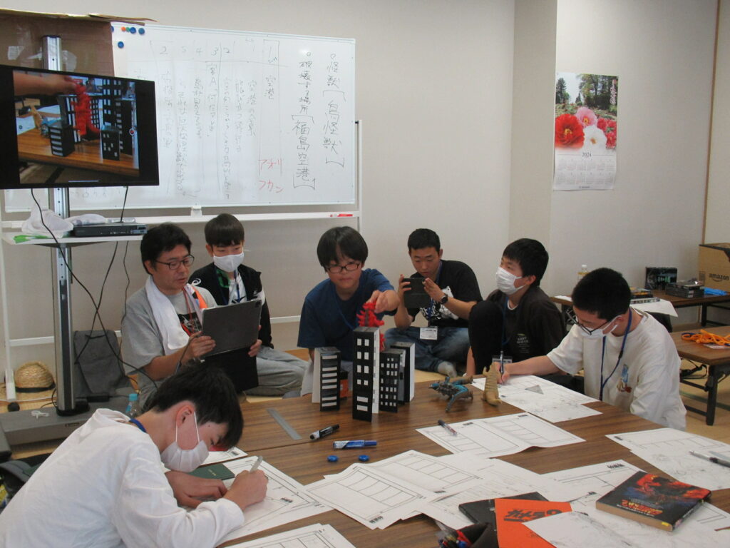 和室の大きな机を囲んで座る講師と塾生が、ホワイトボードに書かれた「怪獣の現れる場所」などの設定案を確認しながら、手元の絵コンテ用紙に熱心にペンを走らせている様子の写真