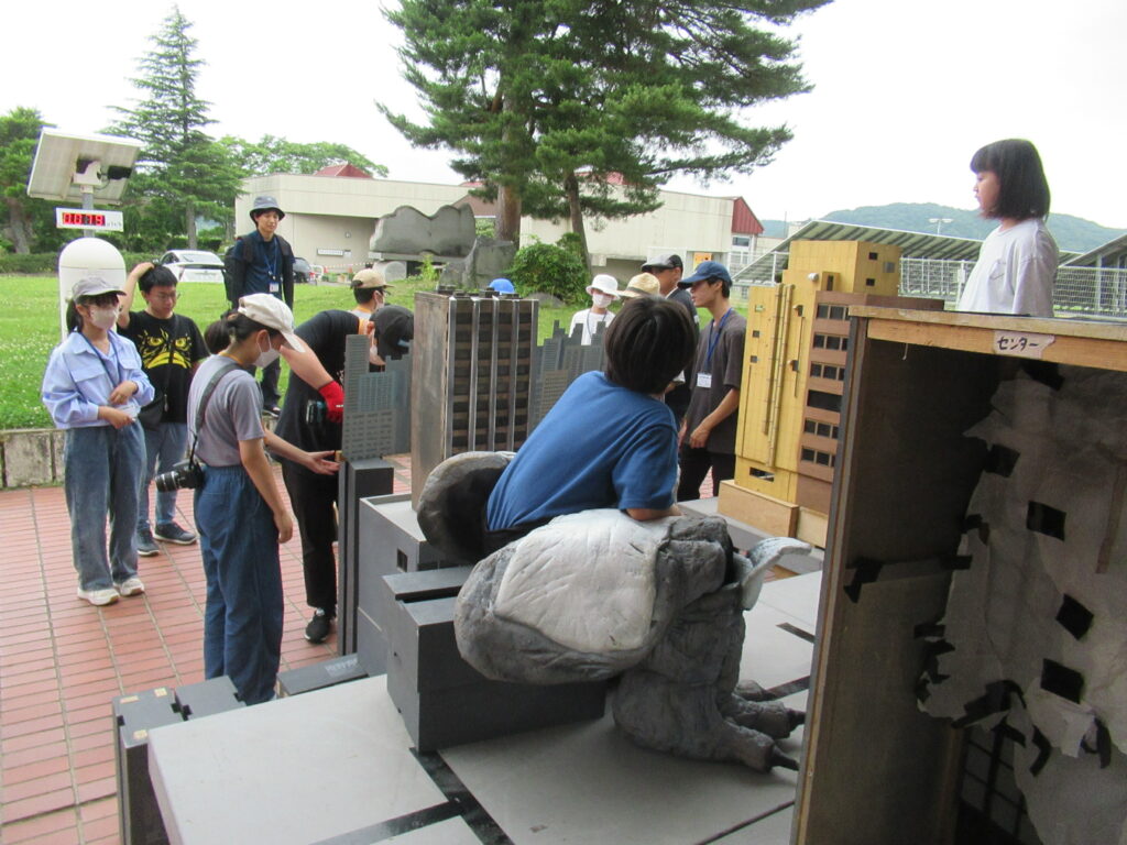 青々とした田園地帯に隣接する屋外セットにて、精巧なビル群のミニチュアに囲まれる中に巨大な怪獣の着ぐるみを着用した男性と、講師や大勢の塾生がカメラやレフ板を手に取り真剣に撮影している写真