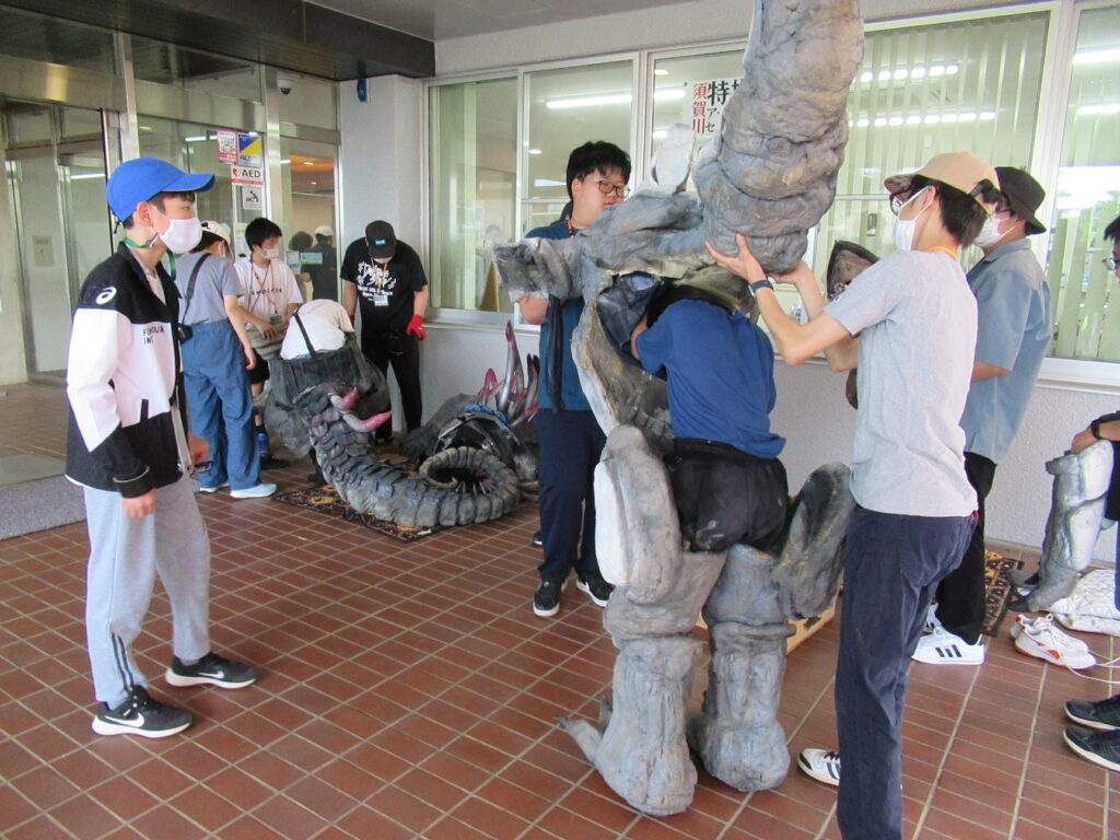 建物の軒下にあるタイル張りのスペースで、数名の塾生が大きな怪獣の着ぐるみを広げ、内部の構造を確認したり実際に足や胴体のパーツを装着したりして準備を進めている写真