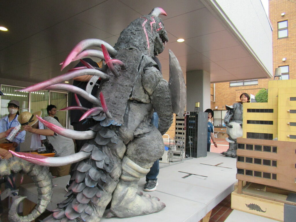 屋外の特撮セットにて、精巧に作られたビル群のミニチュアの背後に巨大な怪獣の着ぐるみが立ち、複数の塾生が動きを真剣に確認している写真