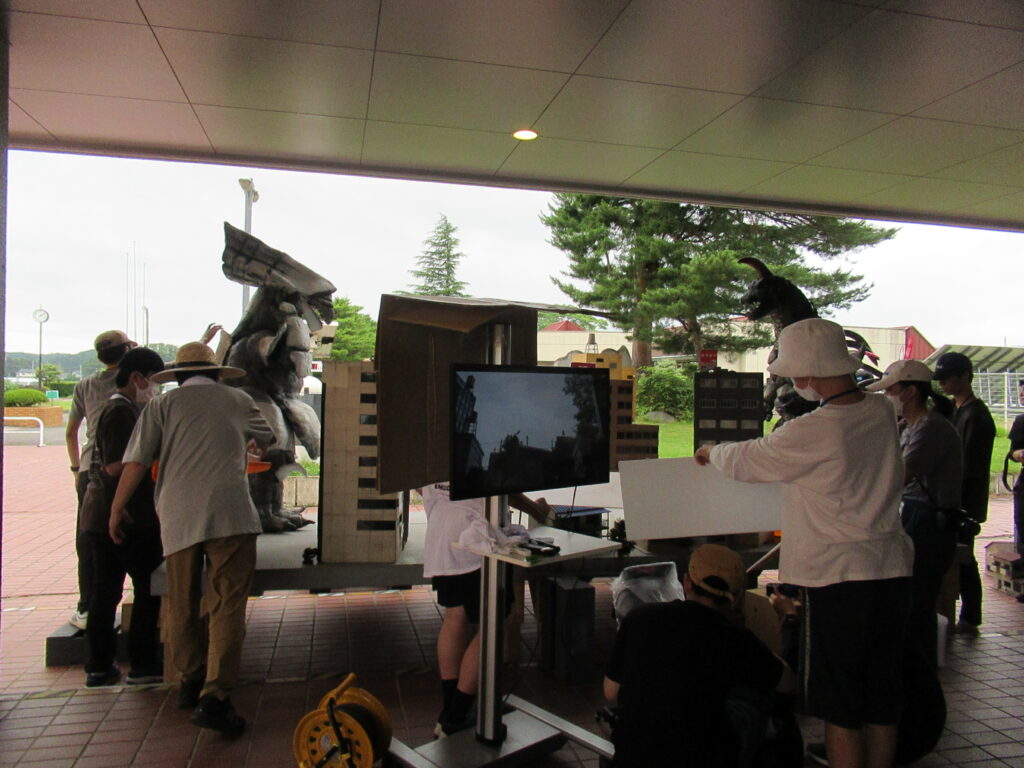 屋外の庇の下で、複数の人々がモニターや白い板を囲み、特撮映画のセットのようなビル群の模型や怪獣のスーツを設営したり調整したりしている、制作現場の風景を捉えた写真