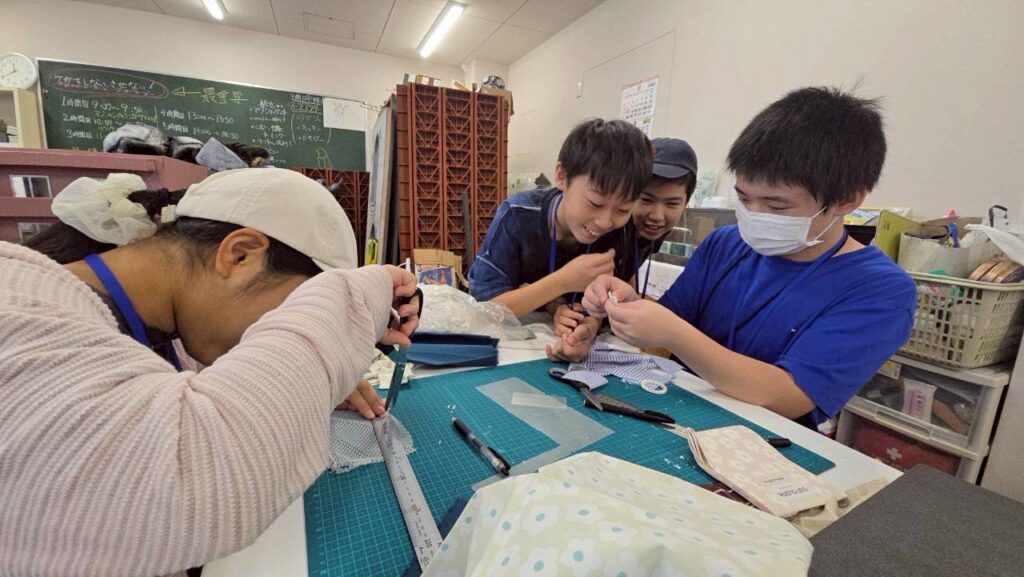 作業台で4人が布を定規で測りカッターや針を使って裁断や縫い合わせの工程を丁寧に進めている様子の写真