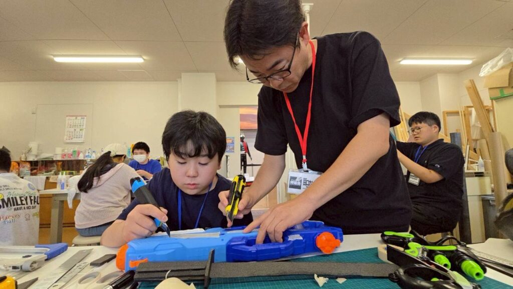 作業台で男性2人が玩具銃を工具で分解し内部構造を確認しながら改造作業を進めている様子の写真