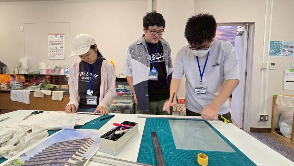 作業台で塾生3人がカッターマット上の素材を定規で測り切り出し作業を丁寧に進めている様子の写真