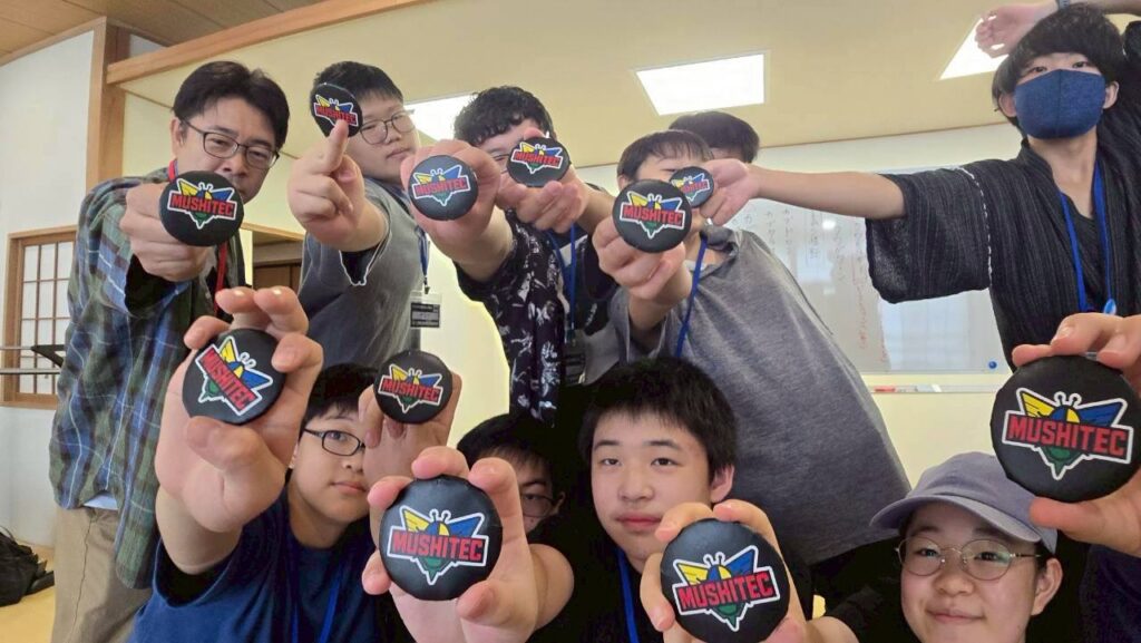 複数の参加者が「MUSHITEC」ロゴのバッジを力強く掲げ前方へ腕を伸ばしてポーズを決める様子の写真