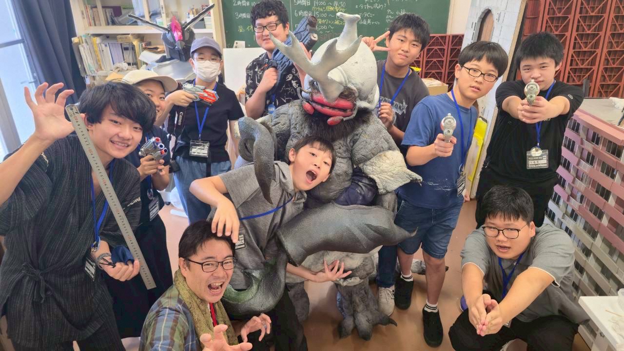 室内で複数の子どもたちが怪獣の着ぐるみを囲んでポーズを取り、手に小道具を持ちながら体験学習や創作活動を楽しむにぎやかな様子が伝わる写真