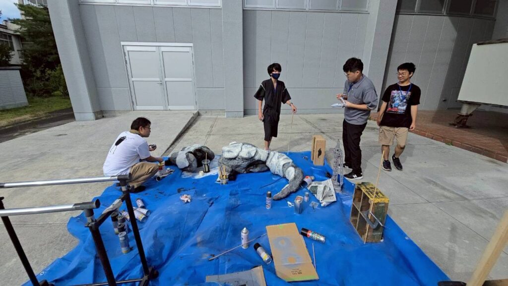 青いシート上で怪獣スーツのパーツを囲み数人が塗装や調整作業を慎重に進めている様子の写真