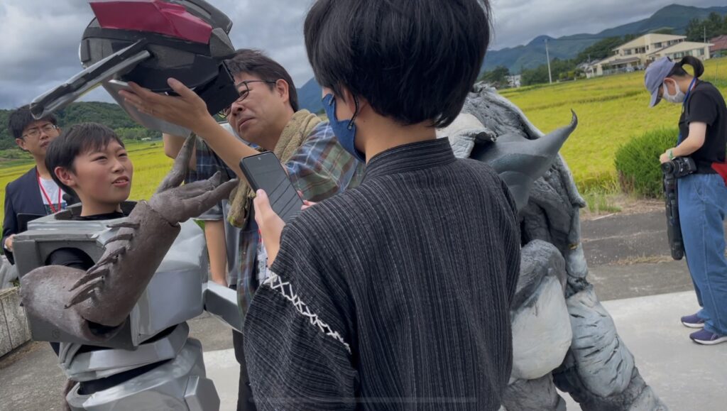 屋外でロボットスーツや怪獣スーツの人物を周囲のスタッフが角度や位置を確認しながら調整作業を進める様子の写真