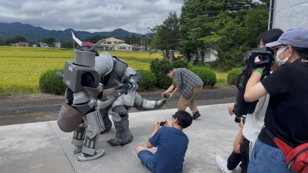 ロボットスーツと怪獣スーツが組み合う動きをスタッフが間近で調整し撮影者たちが構図を整えて記録する様子の写真