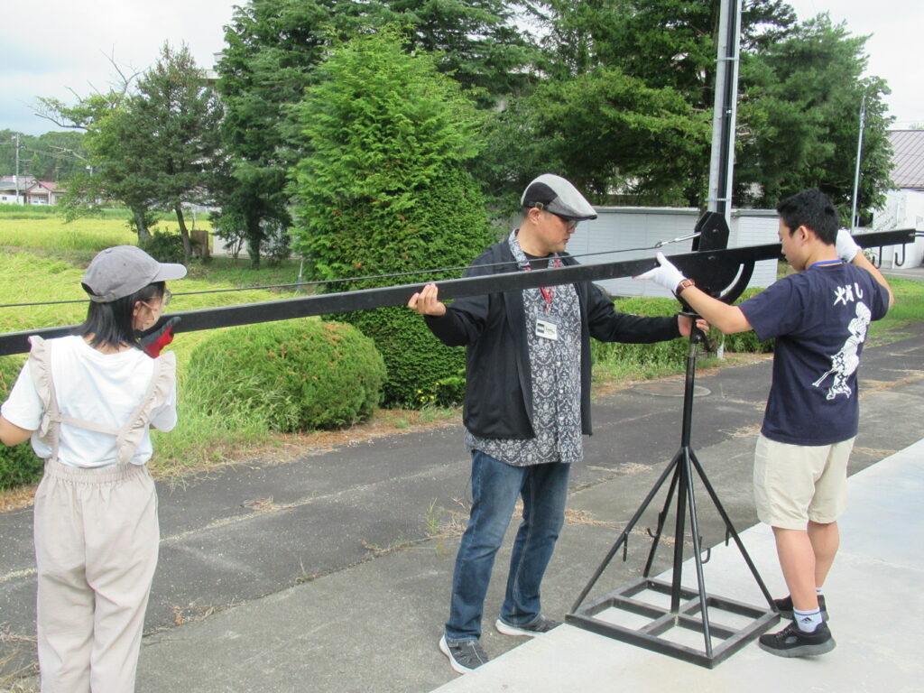 屋外で三人が長い器具を支えるように調整しながら作業手順を確認している様子を捉えた写真