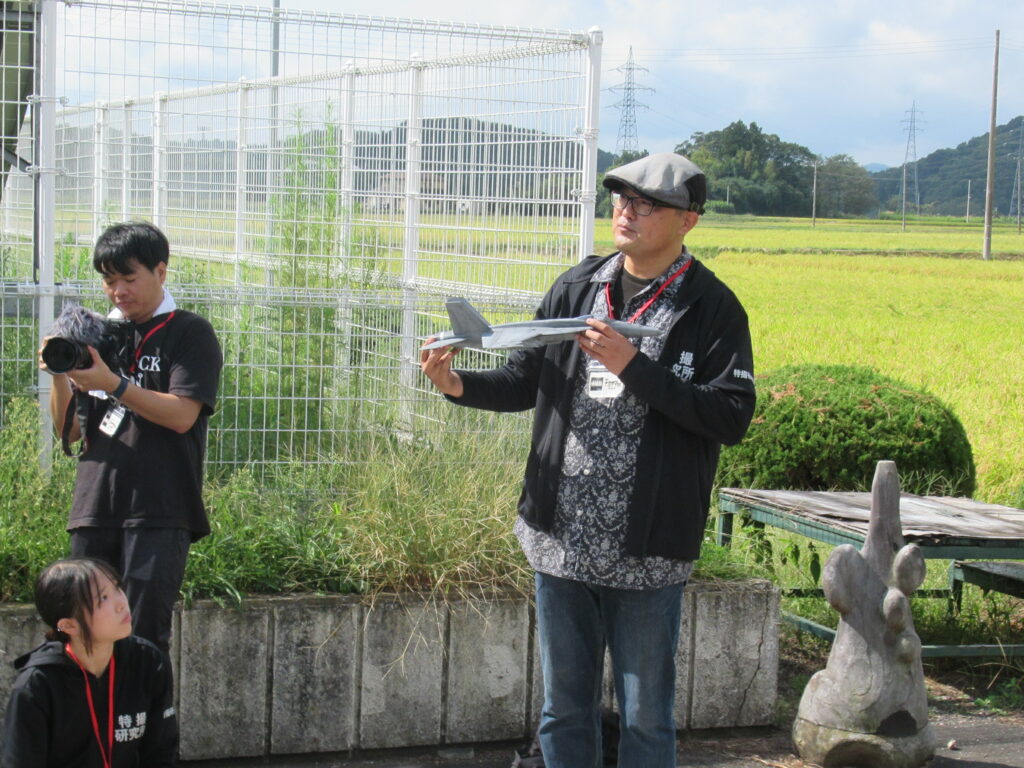 屋外の田園風景を背景に模型の戦闘機を手にする人物と撮影機材を構える参加者が並ぶ写真