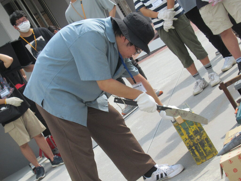 屋外で保護手袋を着けた人物が金属器具で黄色い台を慎重に切断し、その様子を周囲の参加者が見守る写真