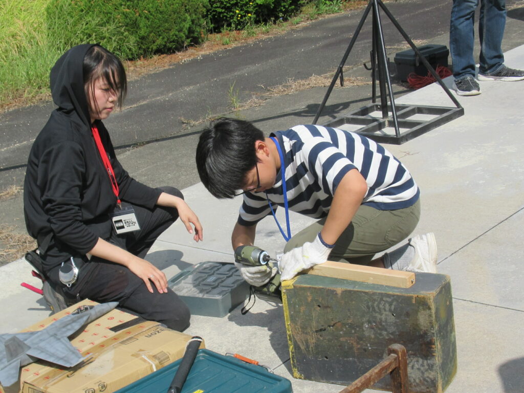 屋外で二人が工具箱を前に金属製の箱を電動ドリルで慎重に加工しながら作業手順を確認する写真