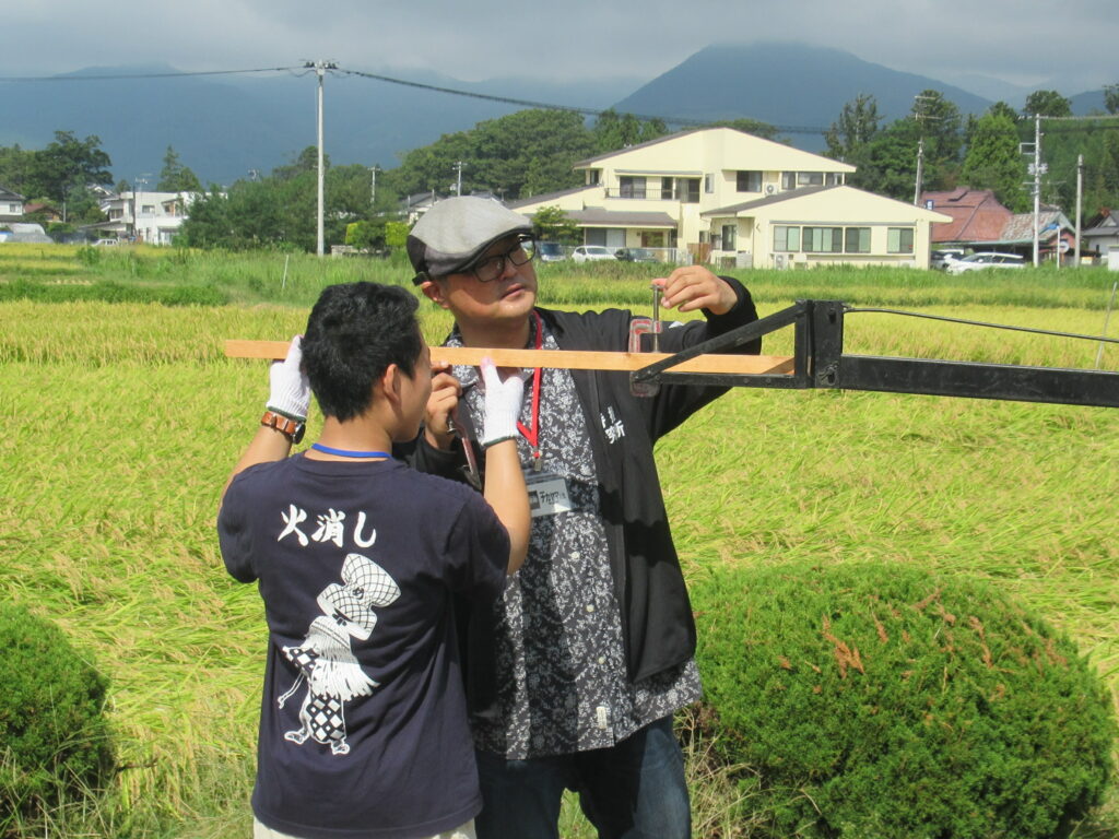 稲穂の広がる屋外で二人が装置に取り付けた木材の位置を細かく調整しながら作業する写真