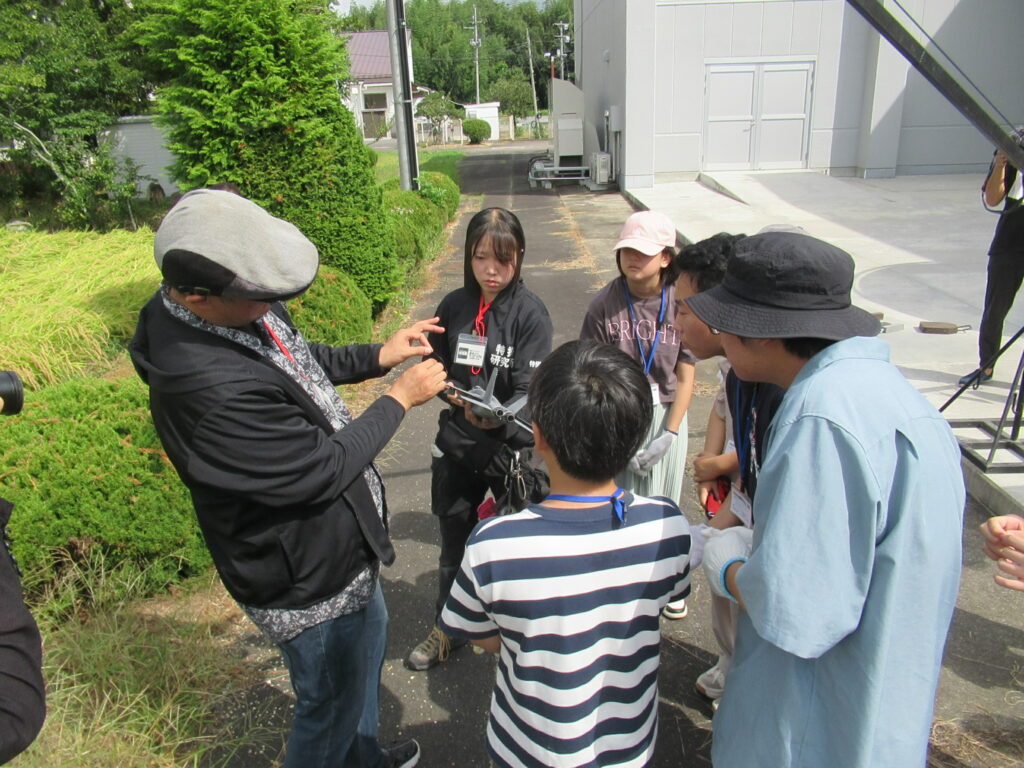 屋外で講師が模型を示しながら周囲の参加者に説明し、皆が身を寄せて確認する様子を捉えた写真