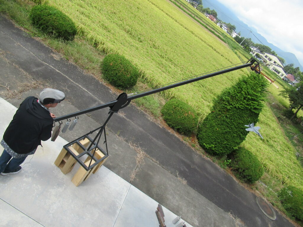 田園風景を背景に長いクレーン装置の先端に模型飛行機が取り付けられ、人物が基部を操作している様子を上方から捉えた写真