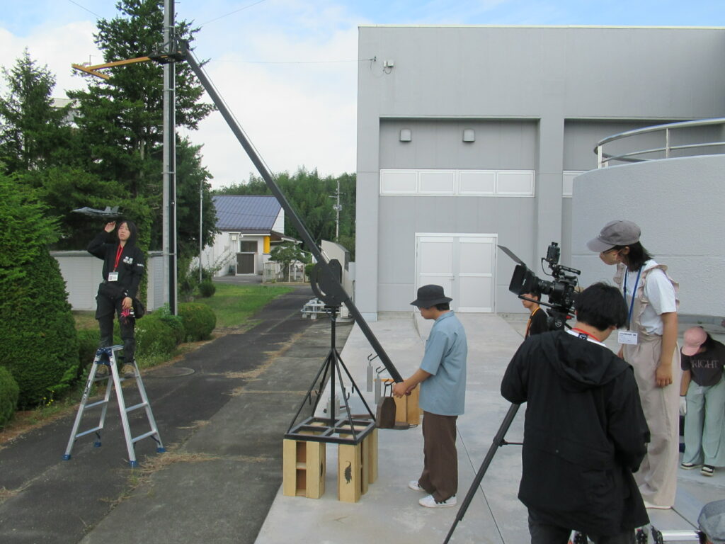 クレーン装置を数人が操作しつつ周囲で脚立やカメラを用いて準備作業を進める屋外の様子を捉えた写真