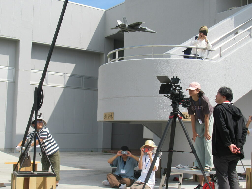 屋外の建物前でクレーンから吊られた模型戦闘機をカメラ機材や見上げる参加者が撮影する様子を捉えた写真