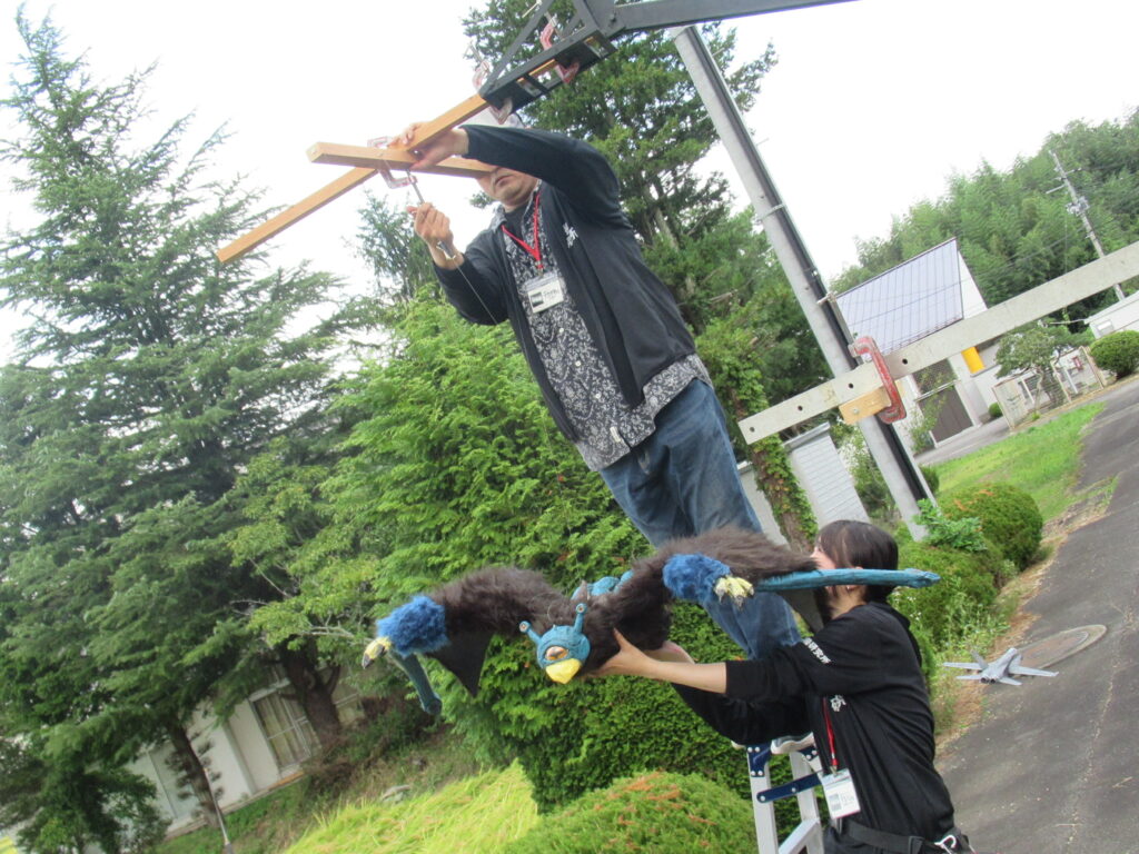 屋外でクレーン装置に木枠を取り付ける人物と、その下で大型の青い怪獣風パペットを支える人物が連携して作業する写真