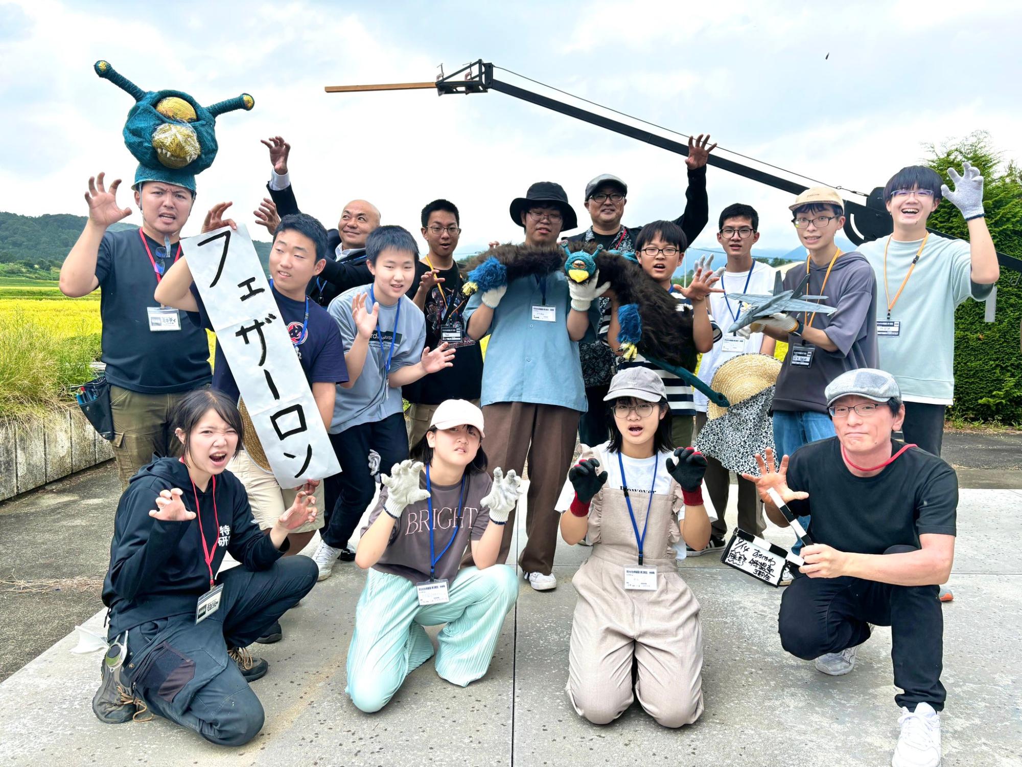 屋外で衣装や小道具を身に着けた大人数の参加者が看板を掲げつつポーズを決めて並ぶ活気ある集合写真