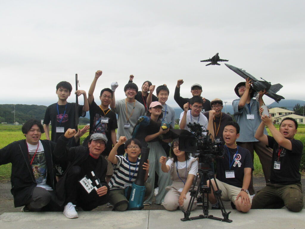 屋外の田園風景を背に大勢の参加者が拳を高く上げたり大型の模型飛行機を掲げたりしながら意気揚々と集合して撮影する写真