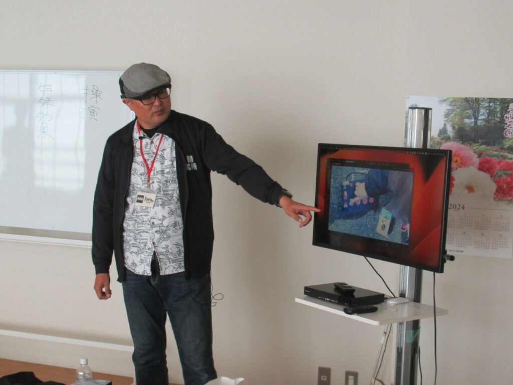 室内で講師がモニターに映る作業映像を指し示し、白板やカレンダーのある教室で解説を行う様子を捉えた写真