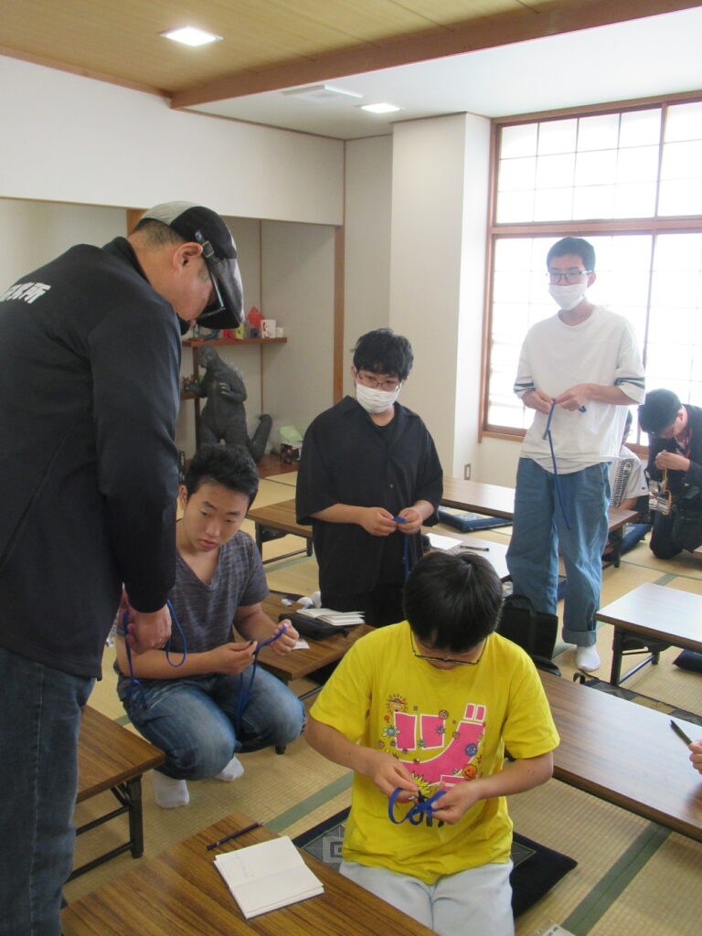畳の部屋で参加者が細長い紐の作業に集中し、講師が机越しに手元を確認しながら指導する写真