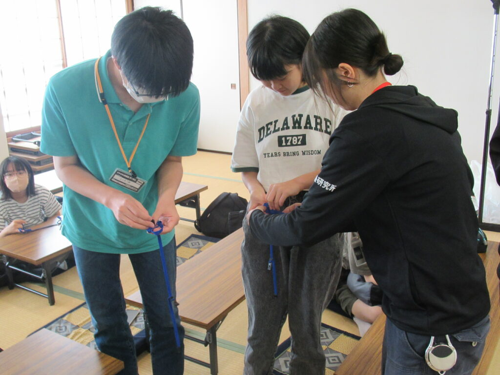 畳の部屋で三人が細い紐を手に取り結び方を確認し合いながら机の間で作業する写真