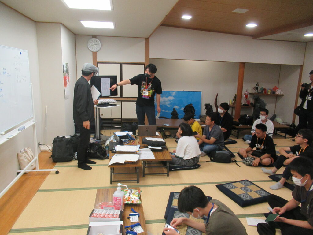 畳の部屋で講師が前方のモニターを指し示し、参加者たちが資料を手元に置いて説明を聞いている様子を描いた写真