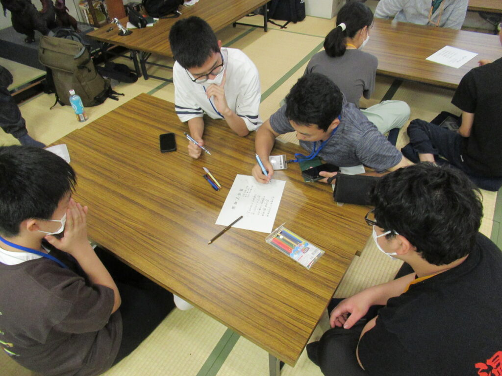 畳の部屋で五人が低い机を囲み、紙に書かれた課題を見ながら意見を出し合い筆記する様子を捉えた写真