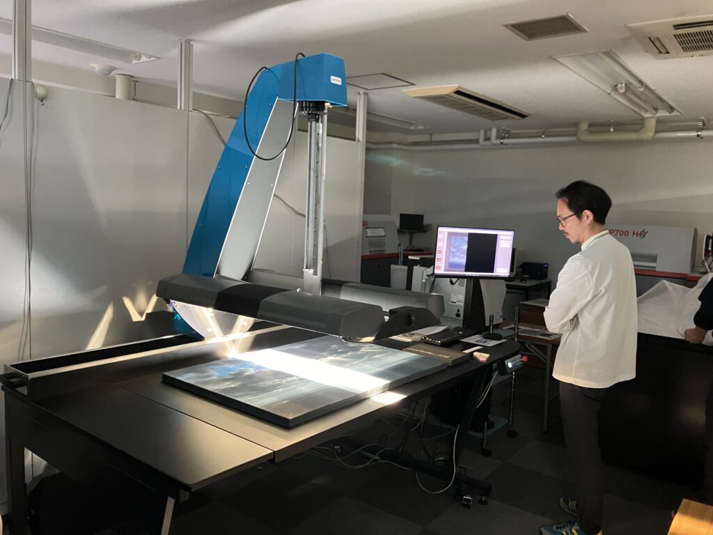 明るい室内で、大型のスキャナーを用いて青空が描かれた精緻な背景画をデジタルデータとして読み込んでおり、傍らに立つ男性がモニターを確認しながら画像の細部や色調の再現性を慎重にチェックしている写真