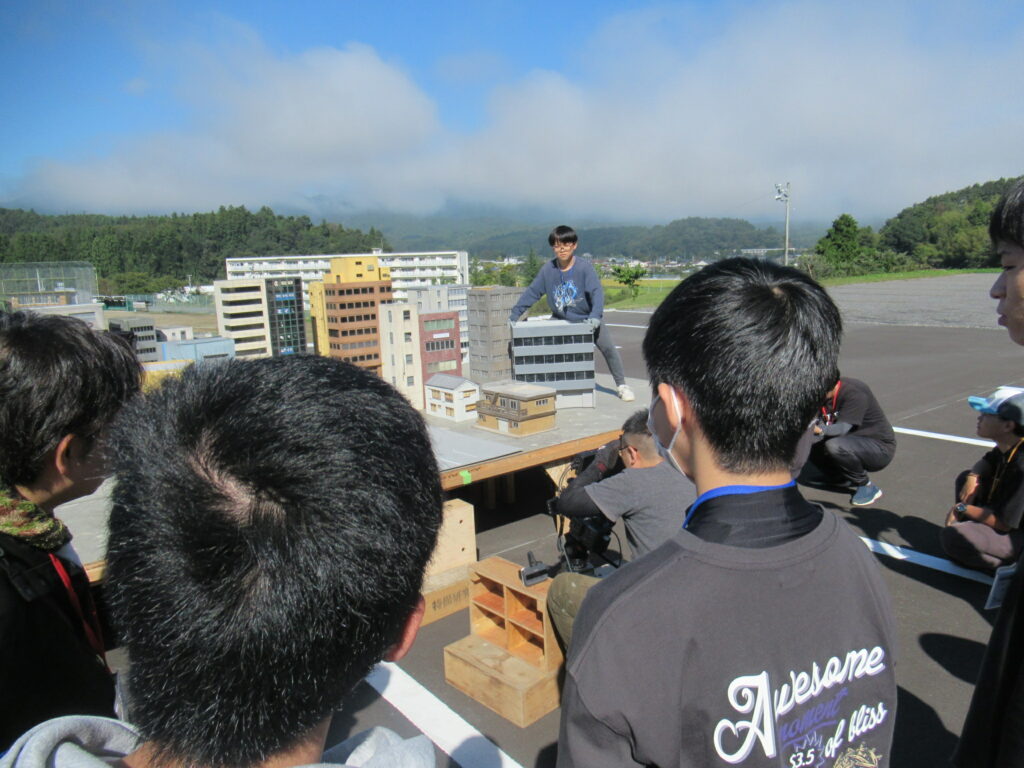 青空の下、屋外のテーブルに並べられたビルや家屋の精巧なミニチュア模型を、数人の若者たちが囲んで撮影や調整を行っている、特撮映画の制作現場のような活気ある風景を捉えた、奥行きを感じさせる構図の写真