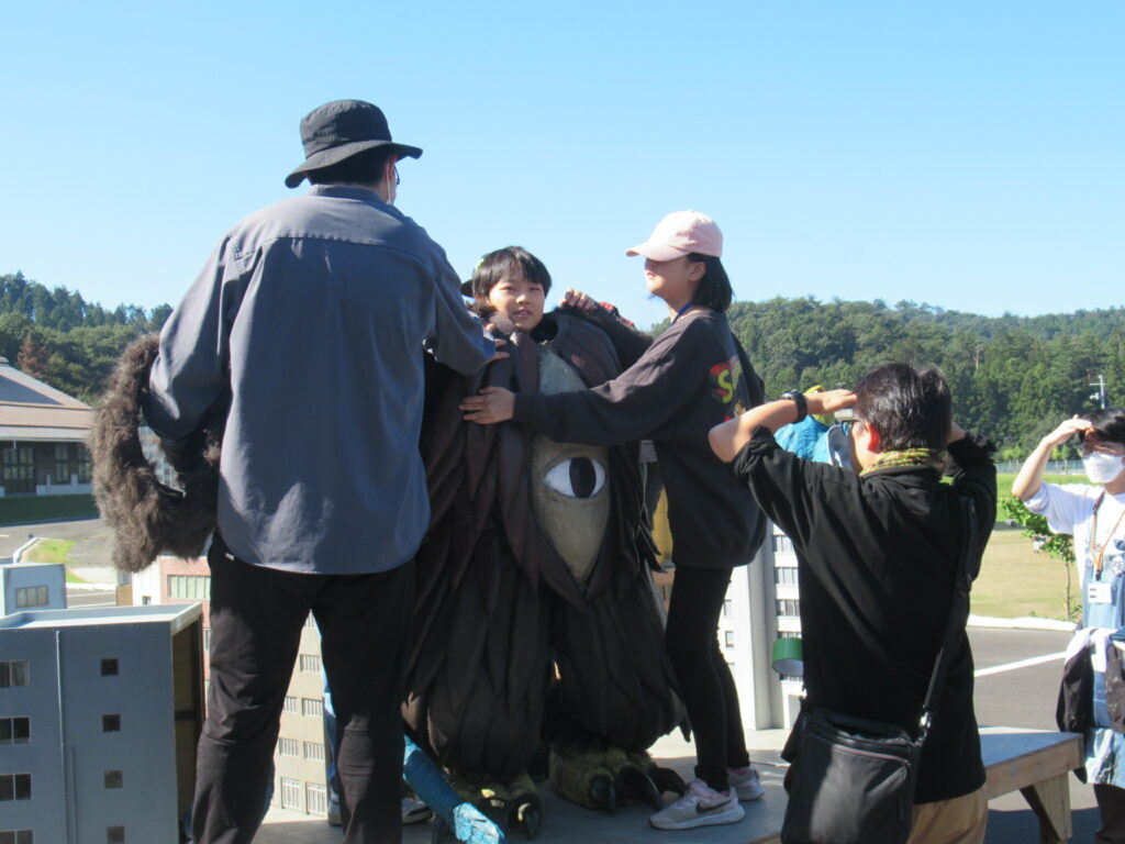 晴天の屋外にて、大きな目が特徴的な怪獣の着ぐるみを着用する少年を周囲のスタッフたちが手伝っており、ミニチュアのビルが並ぶ撮影セットの中で特撮映画の制作風景を切り取ったような臨場感のある写真