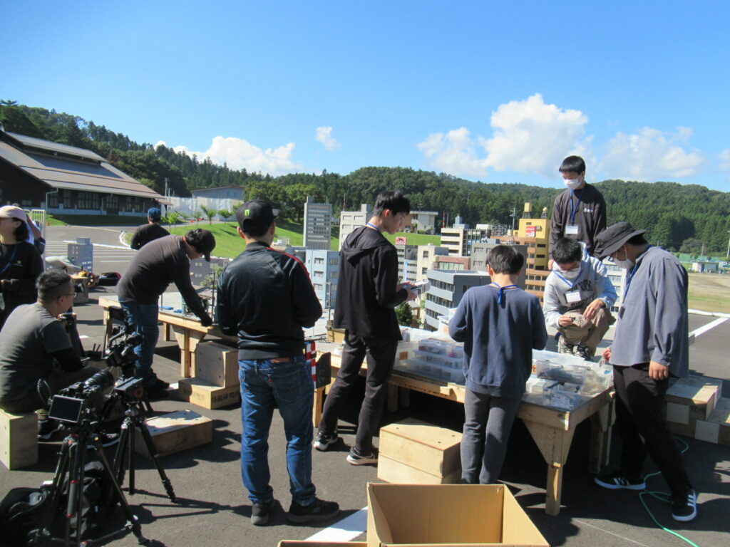 屋外で参加者たちがミニチュアの街並みセットを囲み撮影準備として配置作業を進めている様子の写真